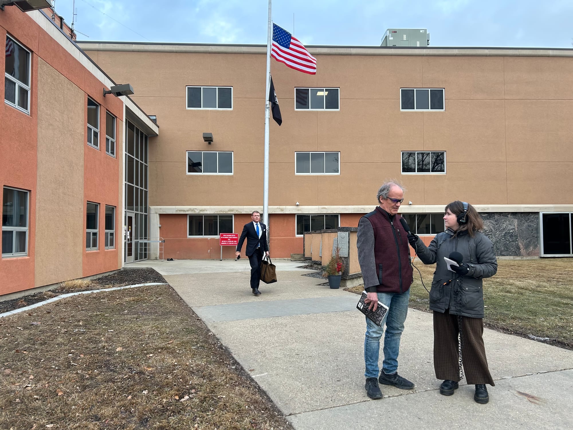 A journalist holds a microphone so a man can speak into it while a man in a suit walks towards them