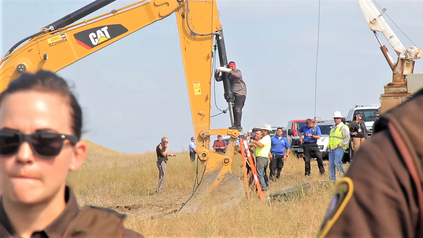 A man stands on a backhoe's arm and attaches his arms to it in protest