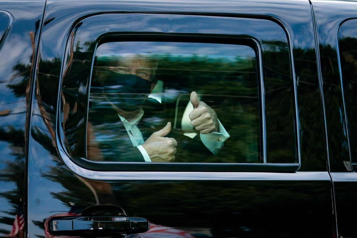 Donald Trump, seated in the backseat of a car, giving two thumbs up while wearing a facemask