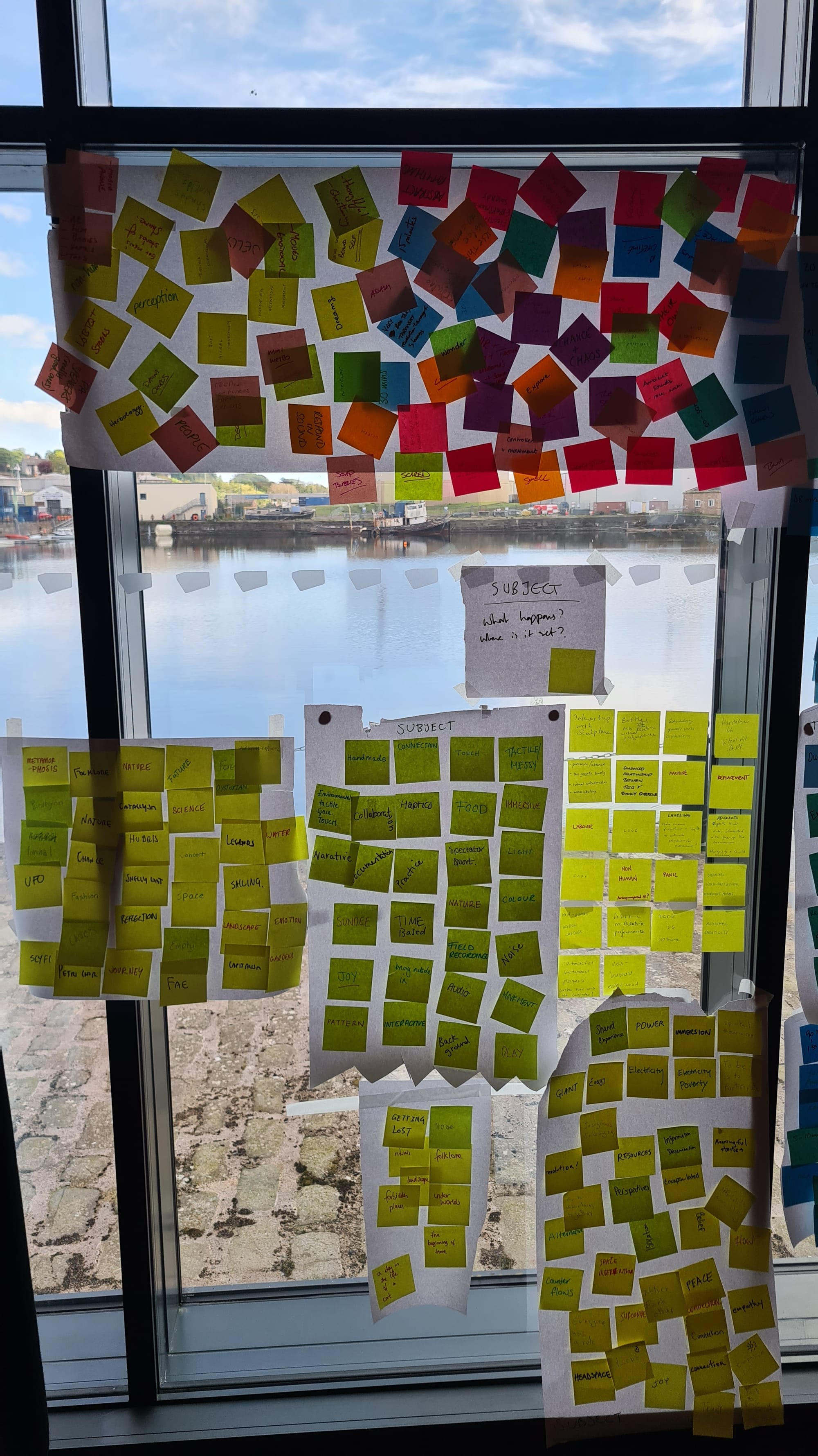 big glass window with sun lit dock and water behind covered in rainbow collage of post it notes with game design jam dompts  on it