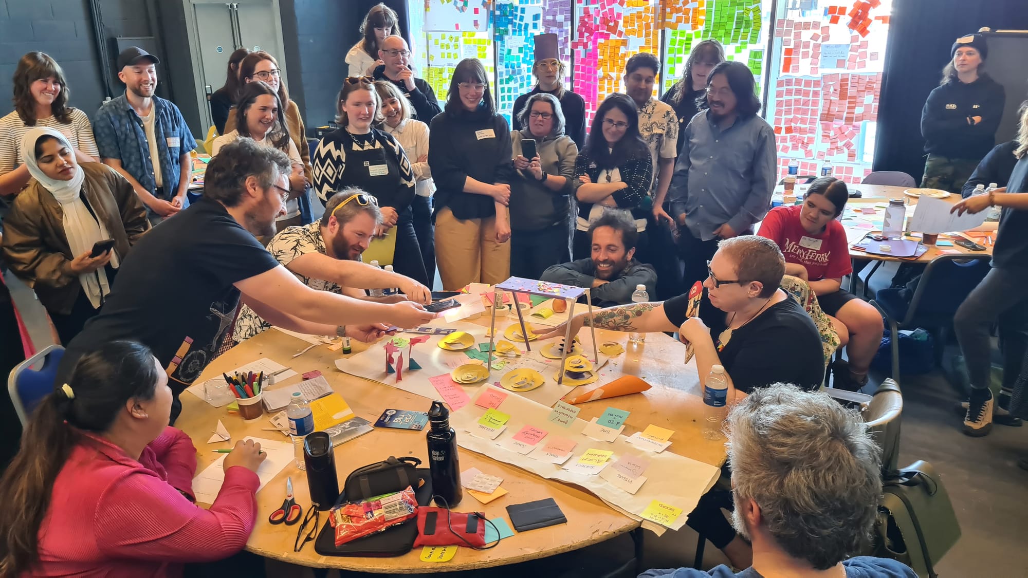 Round table srrounded my mixed crowd of folks standing, some seated around it, a range of wonderful paper craft designed games and related notes and ephemera, muti coloured post it covered windows behind them