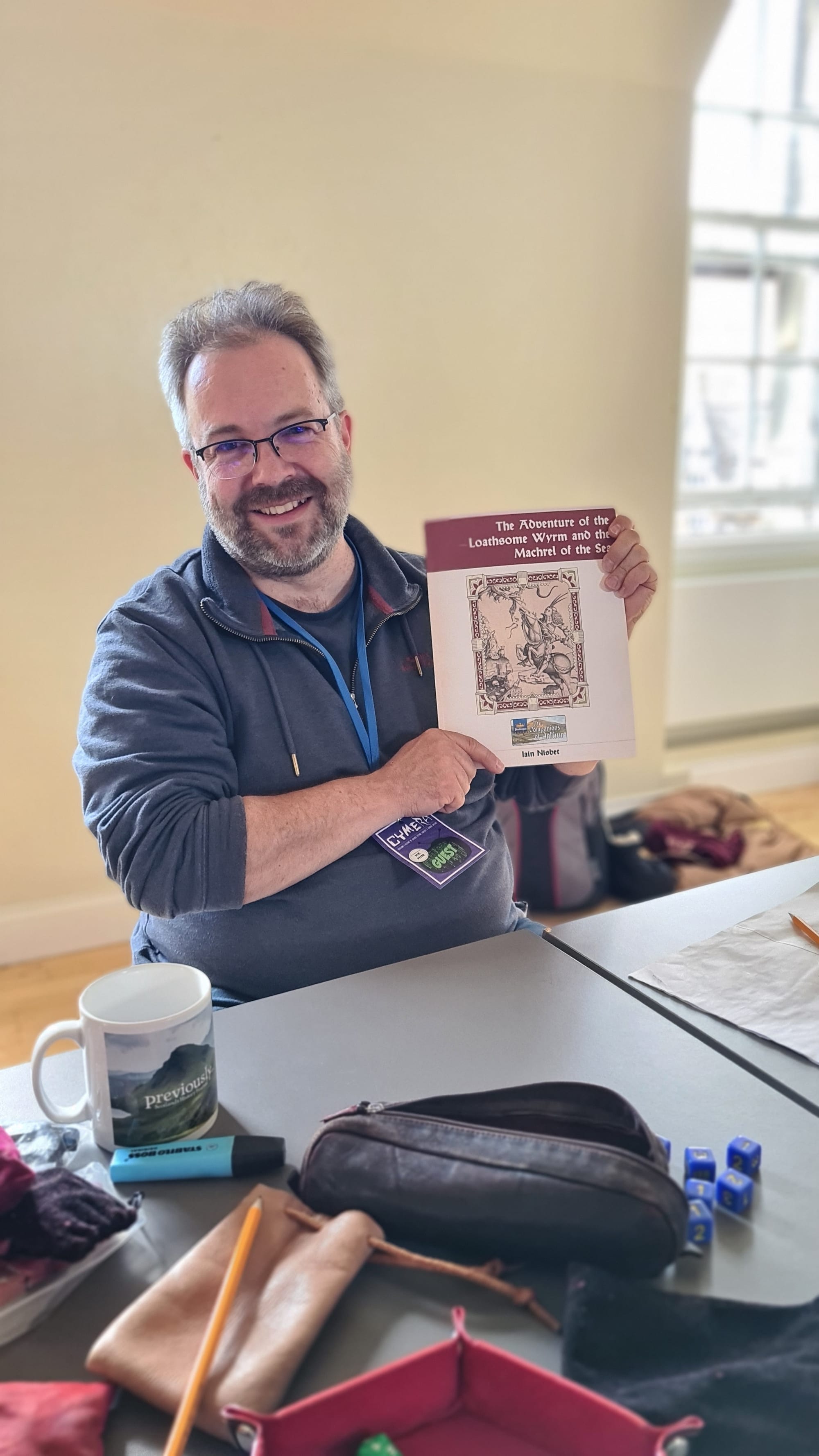 Gent at a table holding up a paper rpg scenario with rpg ephemera (dice etc) on the table