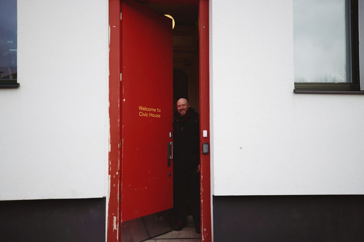 White walled building with black lower wall, tall red door wiht yellow text sayin Welcome to Civic House on it, a smiling blady bearded gentleman is openig the door and looking out