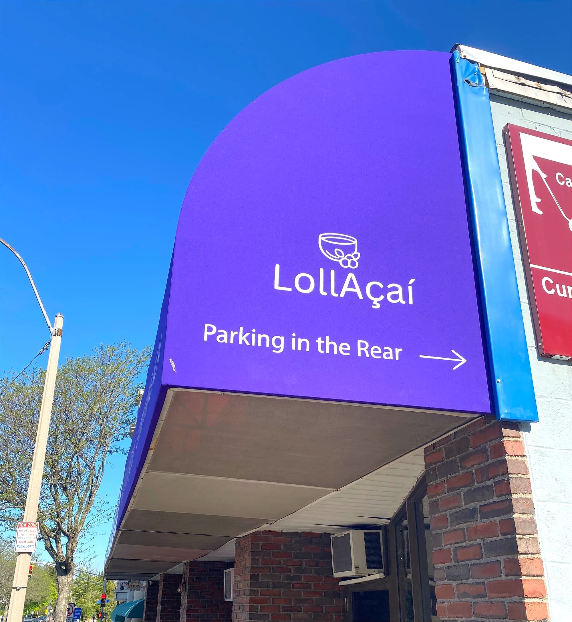 Side view of purple cloth awning against a blue sky. The awning has “LollAçaí” in big letters and “Parking in the Rear” underneath. The building’s red bricks and a red stoplight and green trees are also visible.