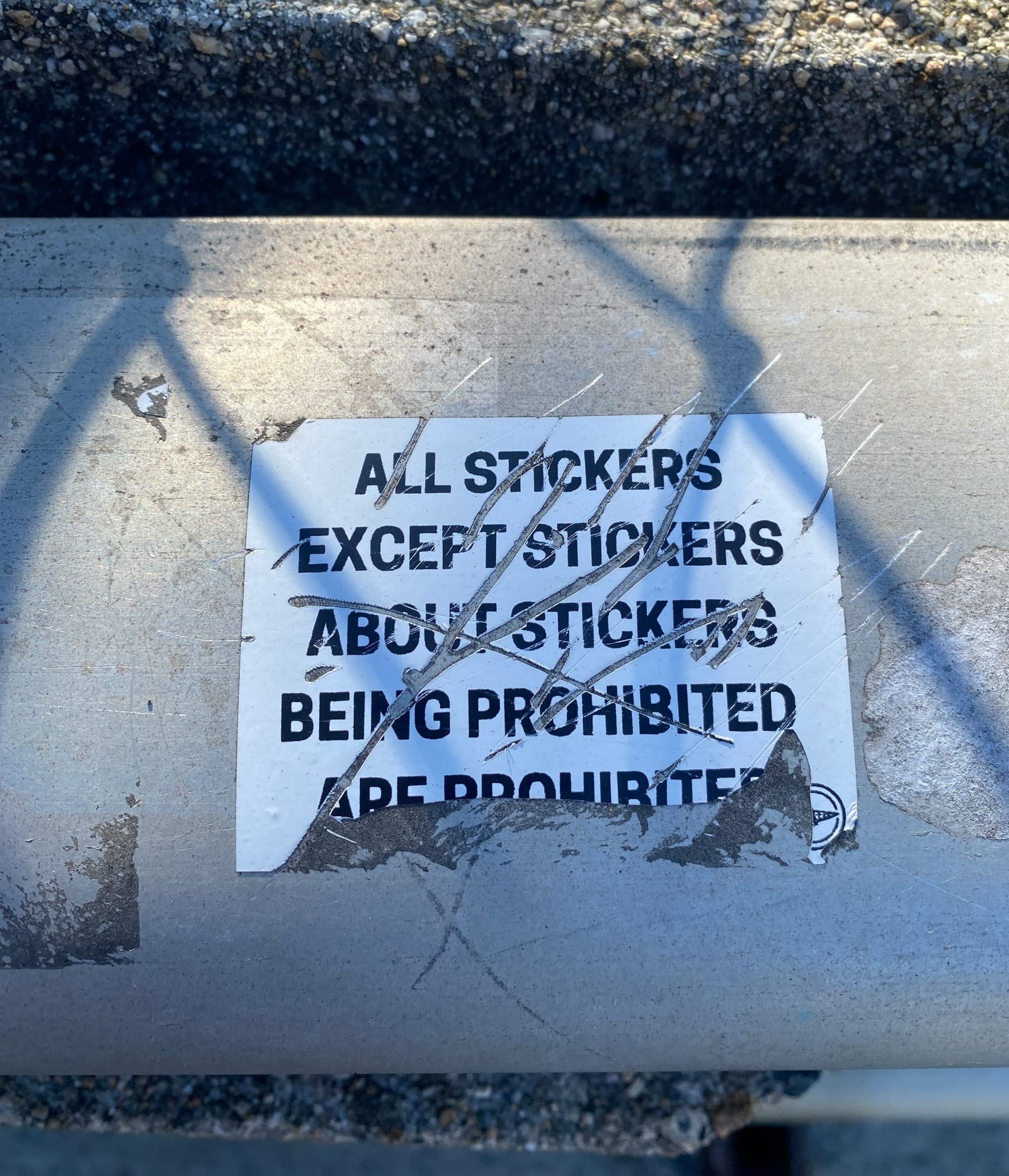 A scratched-up white sticker with black text plastered to a thick metal railing. The text reads, in all caps: “All stickers except stickers about stickers being prohibited are prohibited.”
