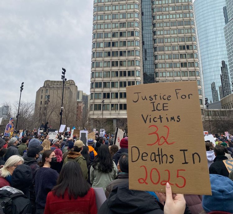 View of hundreds of people bundled against the cold at a rally in Boston. Two skyscrapers in the background. A sign in the foreground reads “Justice For ICE Victims 32 Deaths in 2025.”