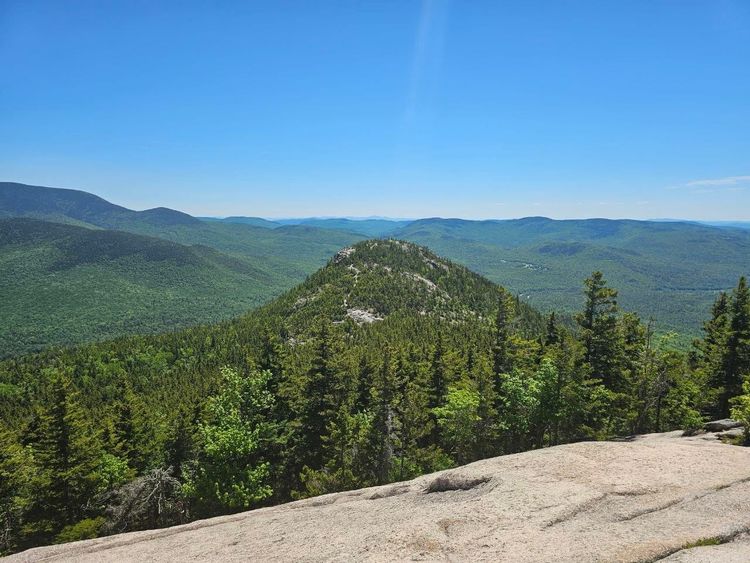 A green, conifer-covered mountain crisscrossed with trails ascends amid a sierra of green and blue peaks in the background, as seen from a rocky slab lookout in the foreground.