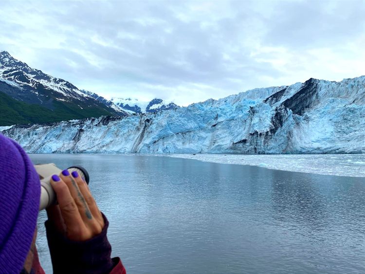 A glowing blue glacier streaked with moraines (dirt & rocks); mountains behind it, ice-strewn water in front. Bottom left: A purple-nailed hand holds binoculars to a purple-capped head.