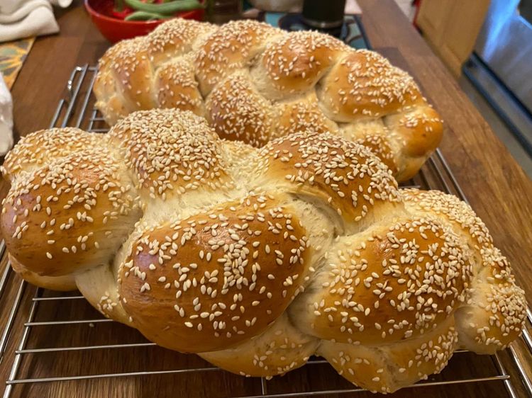 Two braided loaves of challah, browned and sesame-topped, on a cooling rack on a wooden counter.
