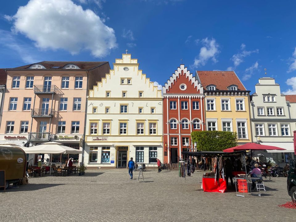 Greifswald's central market square: A row of colorful earth tone buildings with differently shaped roofs; in the foreground, cobblestones, stalls, and umbrellas.
