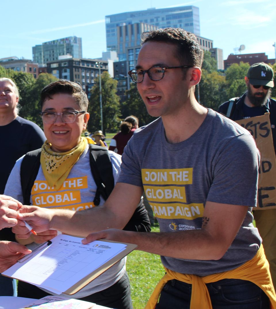Two dark-haired people (I’m one of them) in “Join the Global Campaign” t-shirts holding out a clipboard under sunny skies on the Boston Common.