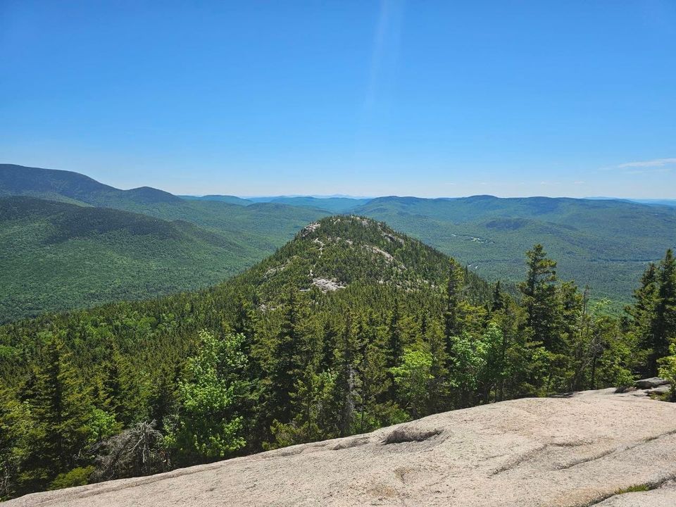 A green, conifer-covered mountain crisscrossed with trails ascends amid a sierra of green and blue peaks in the background, as seen from a rocky slab lookout in the foreground.