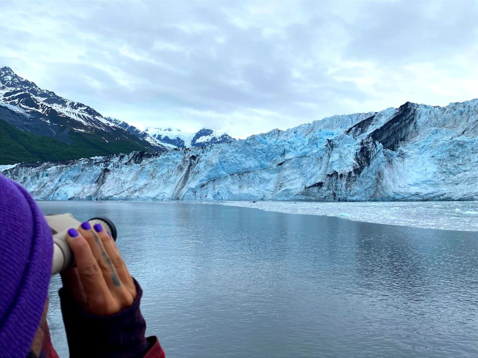 A glowing blue glacier streaked with moraines (dirt & rocks); mountains behind it, ice-strewn water in front. Bottom left: A purple-nailed hand holds binoculars to a purple-capped head.