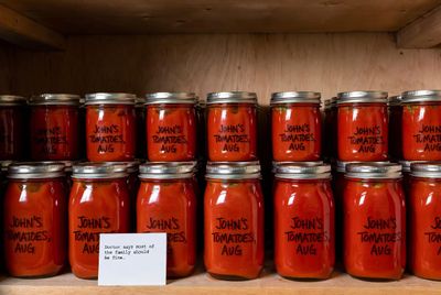 Rows of mason jars filled with canned tomatoes, each hand-labeled "John's Tomatoes, Aug," with a small note card propped in front reading "Doctor says most of the family should be fine."