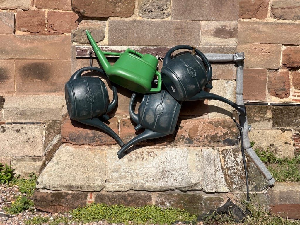 Behind The Photograph: Four Watering Cans Hanging Against An Ancient Wall
