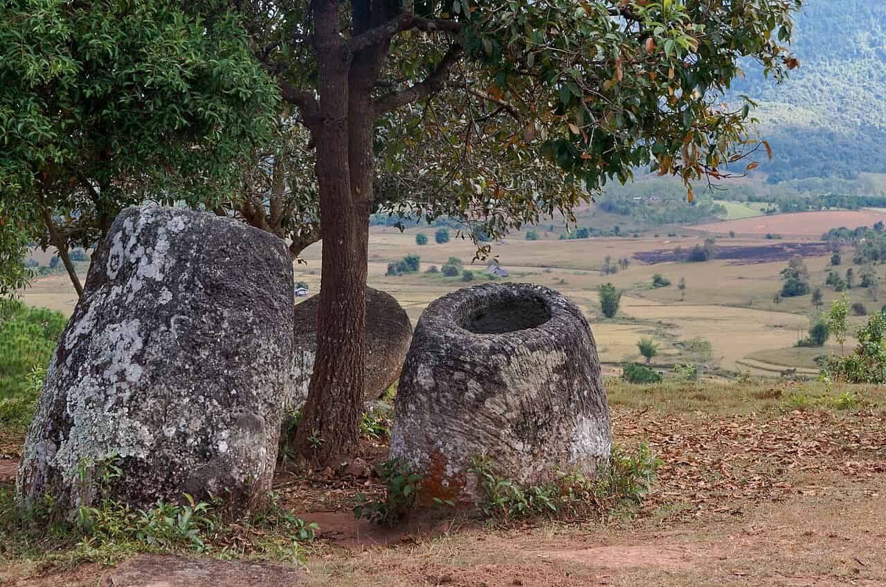 Zwei große, uralte Steinkrüge, etwa einen Meter hoch, stehen auf nackter, rötlich-brauner Erde unter einem Baum an einem Hang. Der rechte Krug steht aufrecht und gibt den Blick auf sein hohles Inneres frei, während der linke geneigt ist. Beide bestehen aus verwittertem, grauem Stein und sind mit Flechten bewachsen. Dahinter erstreckt sich ein weites Tal mit Ackerland und vereinzelten Bäumen bis zum Horizont, grüne Berge ragen empor. Herabgefallenes Laub bedeckt den Boden um die Krüge herum.