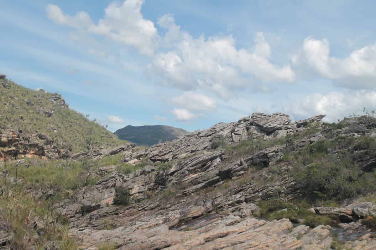 Un paisaje rocoso, sembrado de grietas y maleza. La tierra parece seca bajo el cielo azul claro salpicado de nubes.