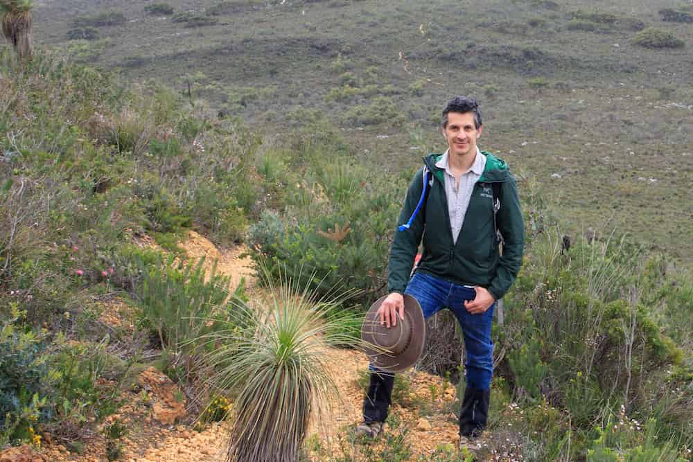 A  person standing on a hiking trail in a mountainous scrubland or fynbos landscape. They are wearing a green jacket, light-colored shirt, blue jeans, and hiking boots, holding a wide-brimmed hat in one hand. The terrain features rocky soil and diverse native vegetation including grassy tufts and small shrubs. In the background stretches a vast hillside covered with similar scrubland vegetation.