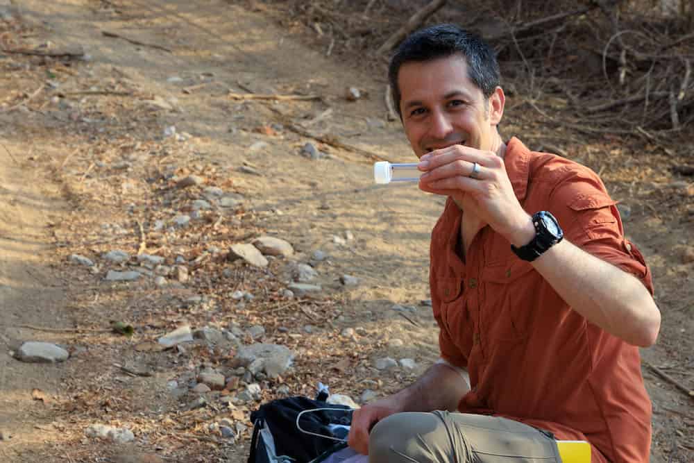 A person in an orange field shirt holding a specimen container while sitting on the ground. They are in a dry, rocky outdoor setting that is a field research site. A dark bag or research equipment is visible beside them.
