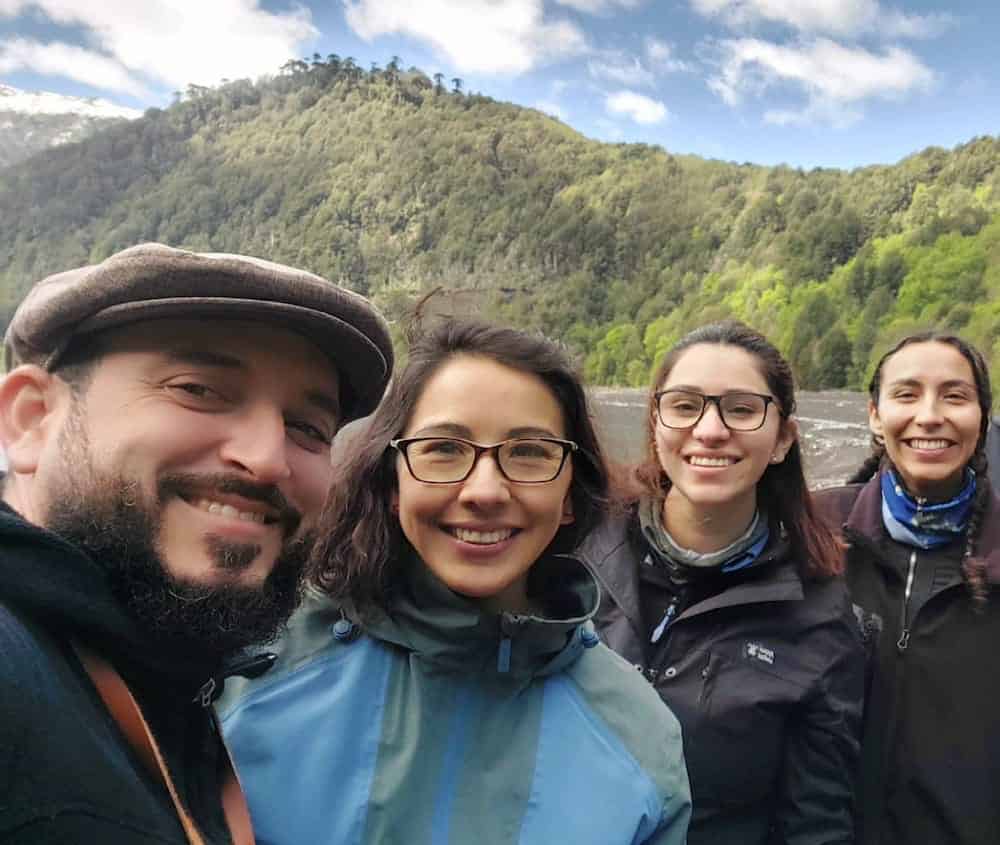 Four researchers smile for a group selfie during fieldwork in a mountainous landscape with forested hills under a cloudy sky. From left: a bearded man in a flat cap and dark jacket, a woman with glasses in a blue and green jacket, and two female students in outdoor gear, representing the collaborative nature of modern lichenological research.