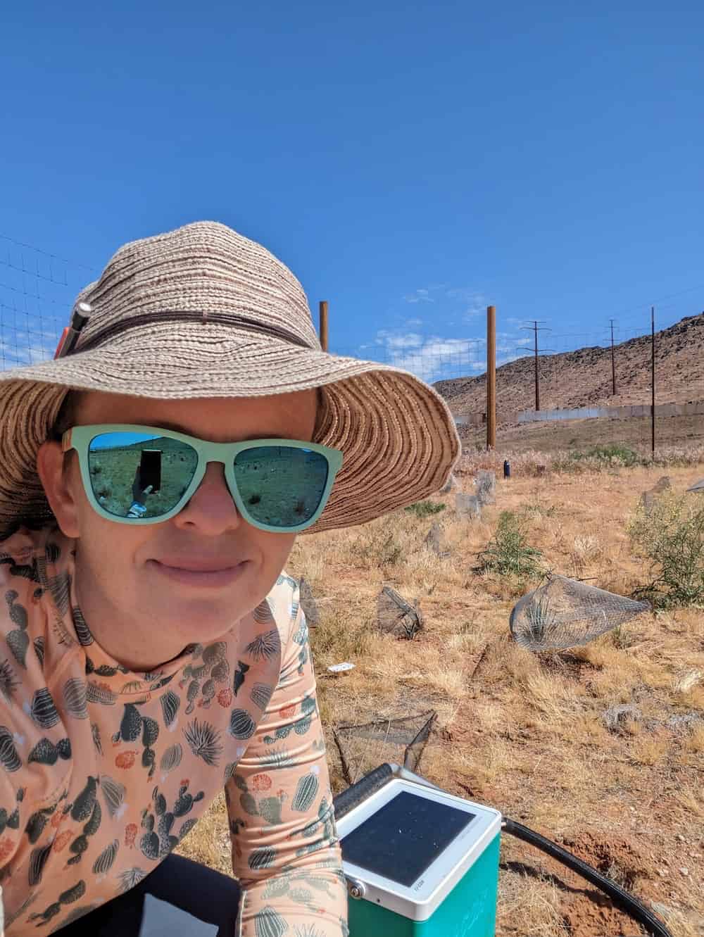 Karolina Heyduk taking a selfie during desert fieldwork, wearing a sun hat and sunglasses with a cactus-patterned shirt, holding scientific measurement equipment, with desert vegetation and research site infrastructure visible in the background under a clear blue sky.