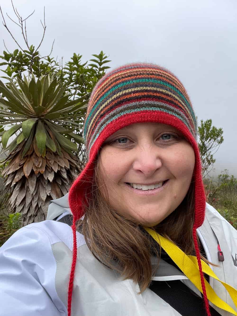 Dr. Laura Lagomarsino smiling at the camera while conducting fieldwork in a tropical environment. She wears a colorful striped knit hat with red trim and a light-colored field jacket with yellow straps. Behind her is a distinctive tropical plant with large, paddle-shaped leaves arranged in a rosette pattern, characteristic of high-altitude Andean vegetation. The misty, cloudy background suggests she is working in cloud forest conditions.