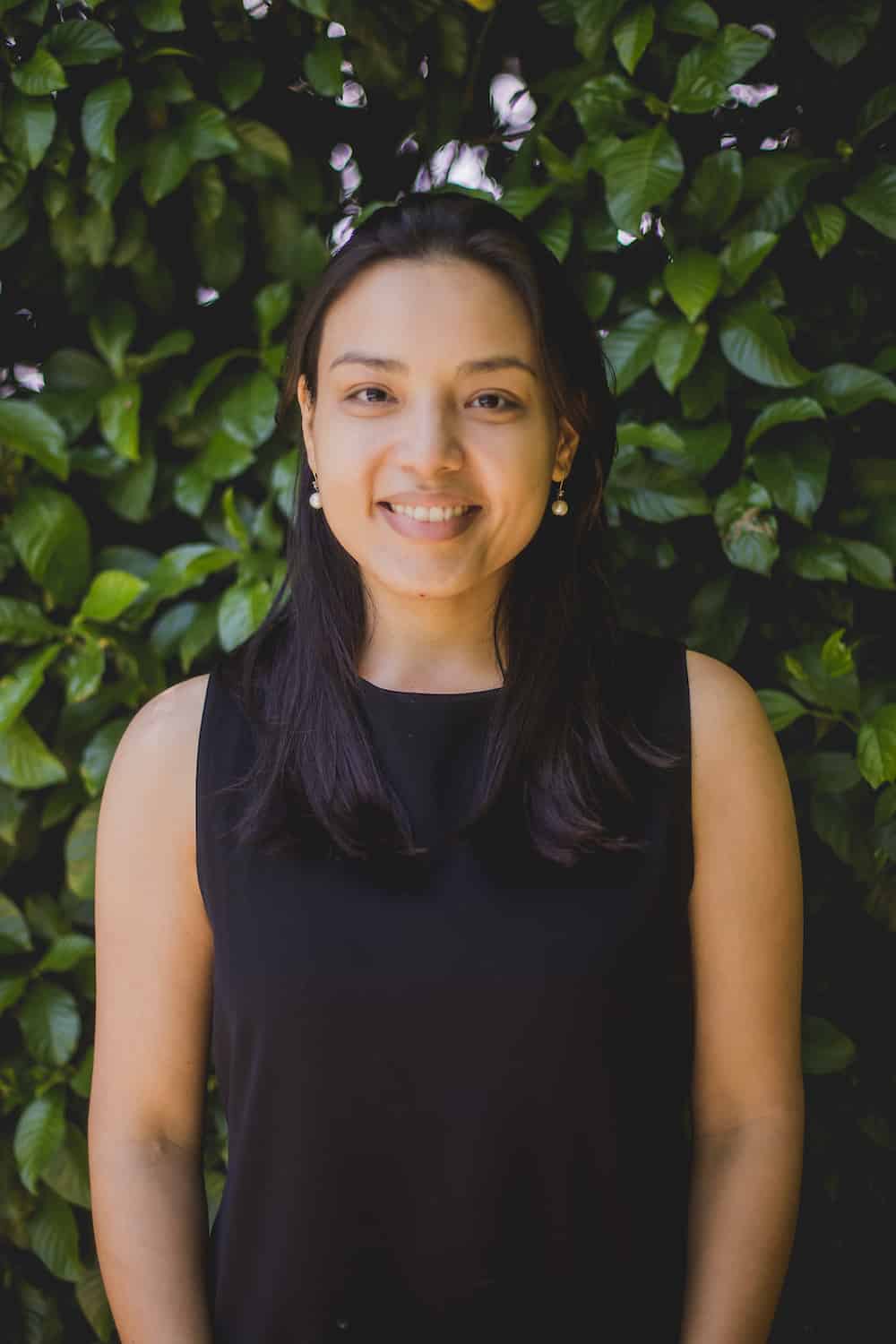 Dr. Juliane Ishida, a smiling woman with long dark hair wearing a black sleeveless top and pearl earrings, photographed against a lush green backdrop of leafy plants with small pink flowers.