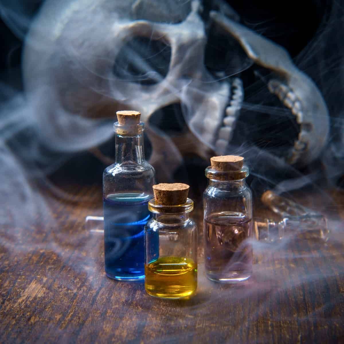 A skull, in front on a table, three bottles of coloured liquid with eldritch smoke around them.