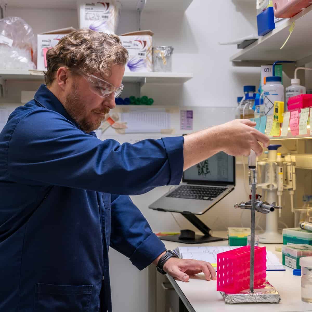 A laboratory researcher wearing safety goggles and a blue lab coat uses a pipette to handle samples in a scientific laborator