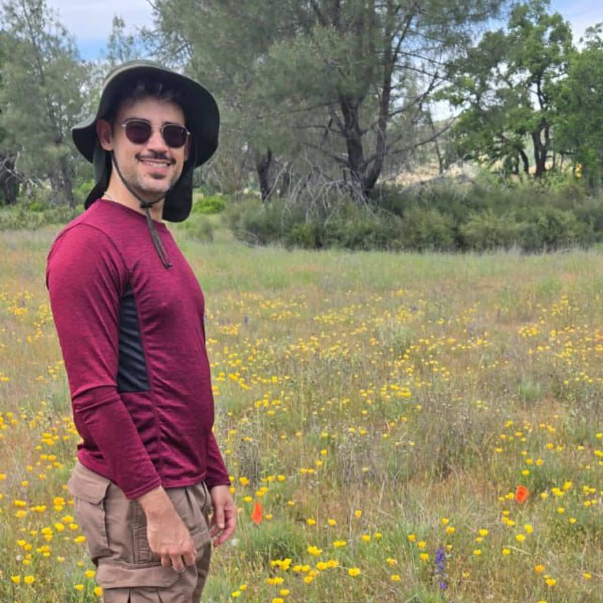 A man in a deep red t-shirt and heavy duty sunhat smiles at the camera. Behind him are many flowers, but how many pollinators