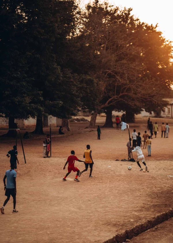 People play football on a dusty and bumpy pitch in the shade of trees.