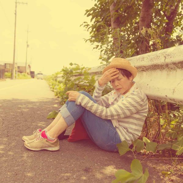 A person in a striped shirt and hat sits on the ground leaning against a concrete wall, with one hand covering their face in