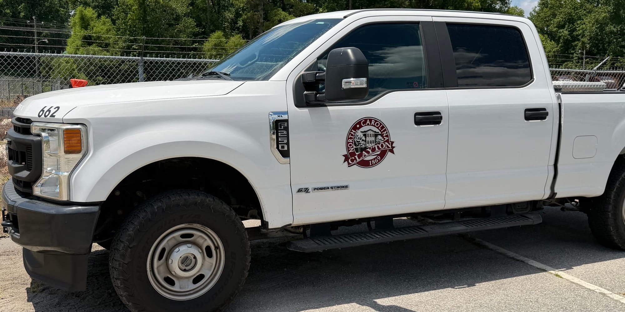 A white Town of Clayton pickup truck parked beside a chain-link fence on a sunny day, with trees and utility infrastructure in the background.