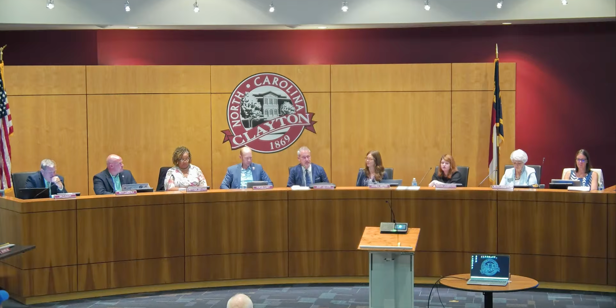 Members of the Clayton Town Council sit behind the dais in the Town Hall council chambers, with the Town of Clayton seal on the wall behind them.