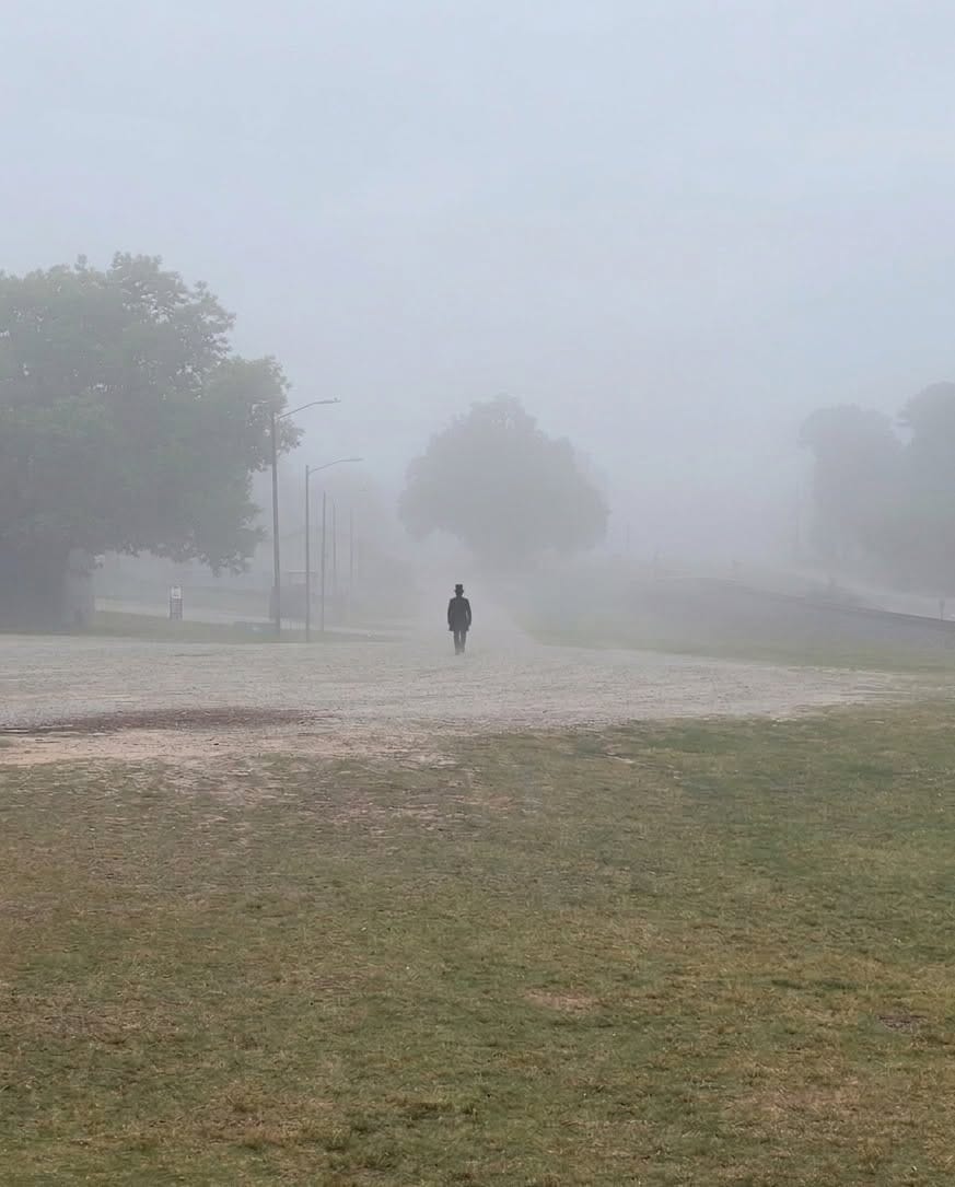 A lone person in dark clothing and a top hat walks away through heavy fog across a grassy downtown area. Trees, streetlights and nearby buildings fade into the mist. The person is identified by the user as Doug Pawlak of Clayton Ghost Walks.