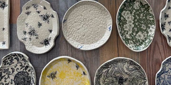 Assortment of handmade ceramic trays and dishes arranged on a wooden floor, featuring floral, botanical, bee and cactus patterns in black, blue, green and yellow.