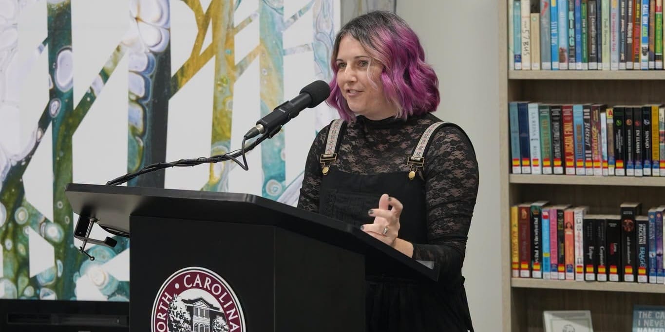 A woman with pink hair speaks at a podium inside Hocutt-Ellington Memorial Library, with bookshelves and a colorful wall mural behind her.