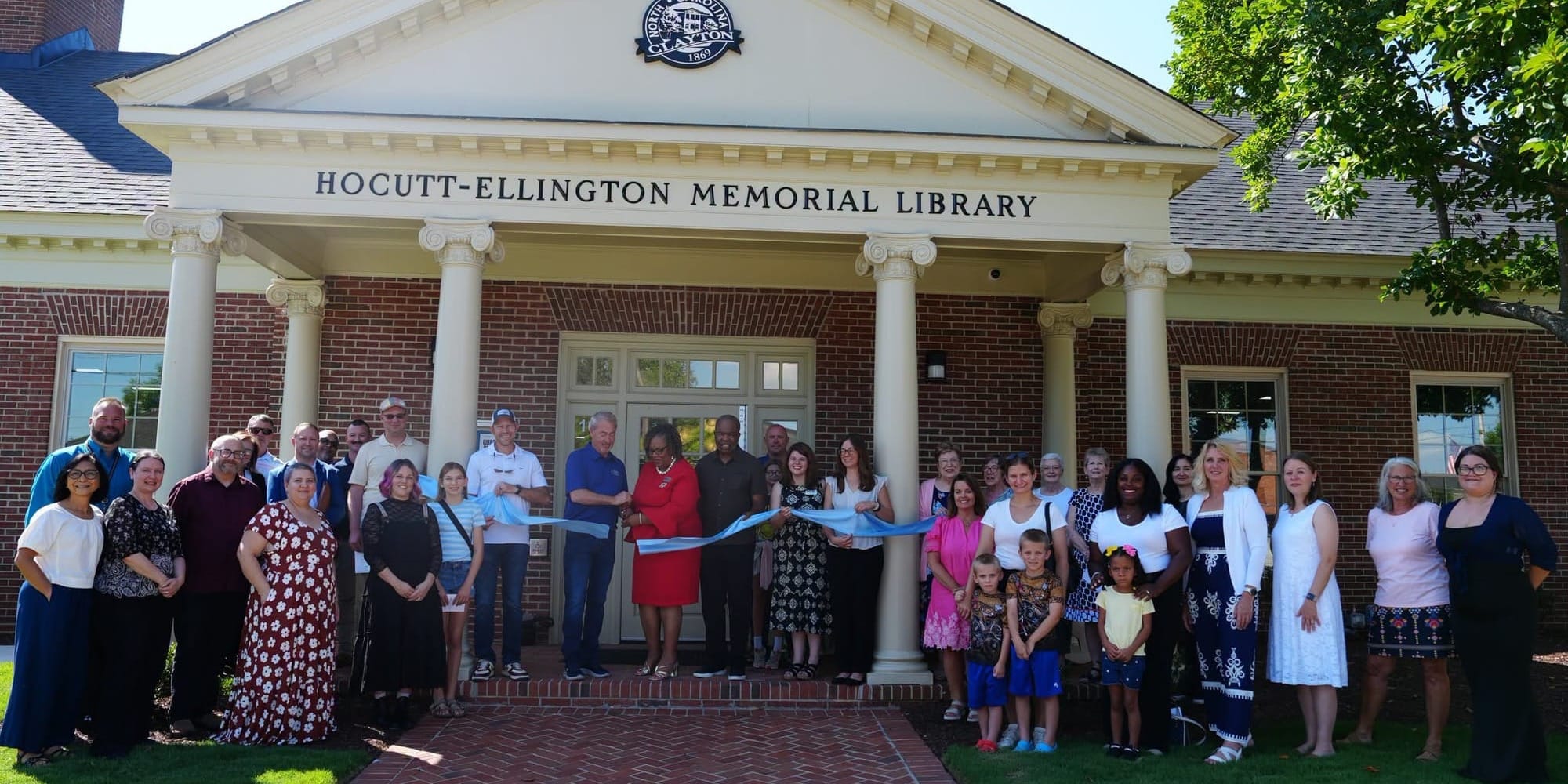 A large group of people stands outside Hocutt-Ellington Memorial Library in Clayton for a ribbon-cutting ceremony, holding a blue ribbon across the front entrance of the brick building.