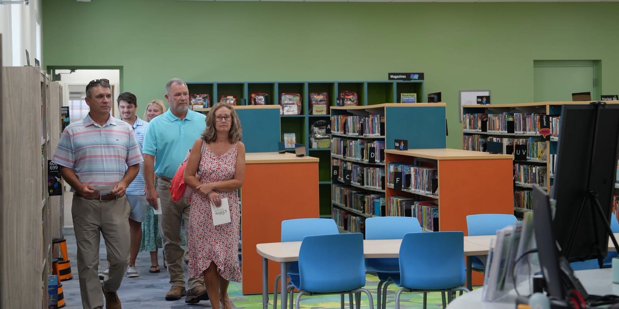 A group of people walks through the renovated Hocutt-Ellington Memorial Library, passing bookshelves, computers, tables, and colorful seating in a bright children’s area.