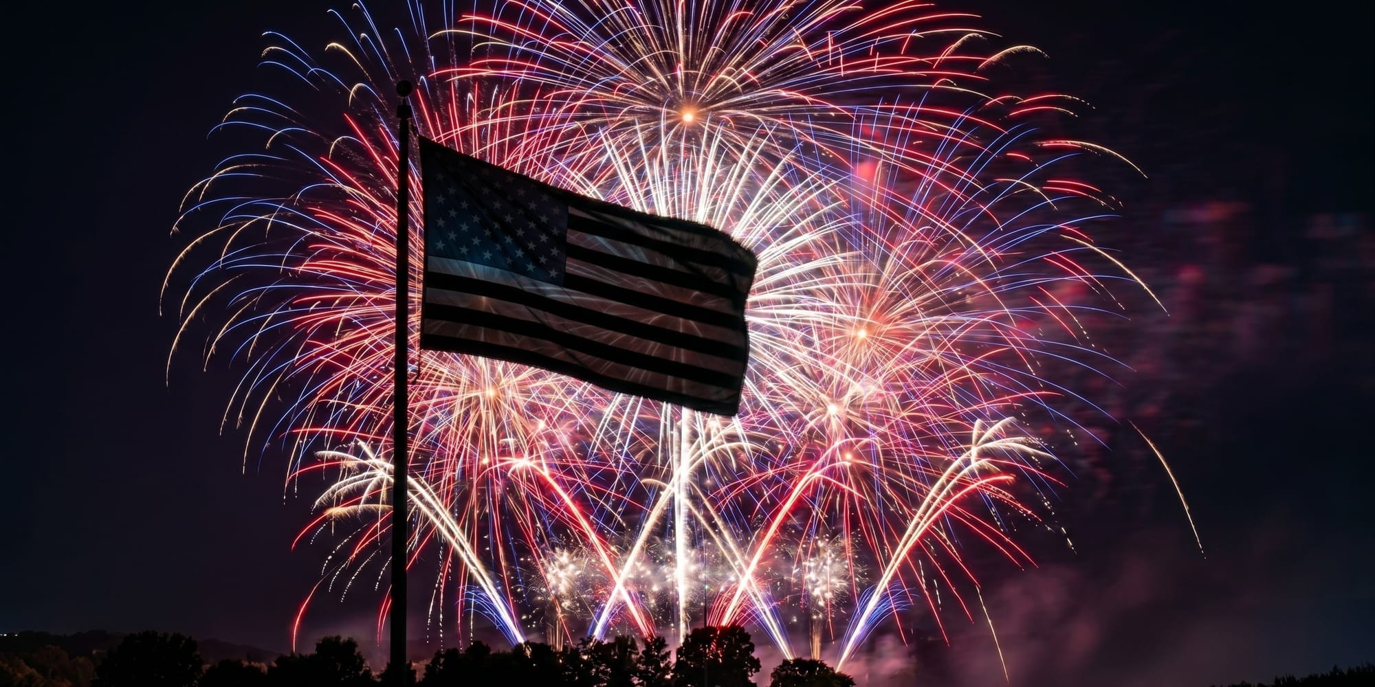 An American flag is silhouetted against red, white, and blue fireworks bursting across the night sky.