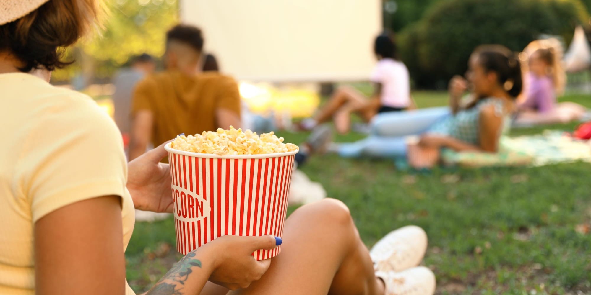 A person holds a large tub of popcorn while sitting on the grass at an outdoor movie screening, with a projector screen and other moviegoers blurred in the background.