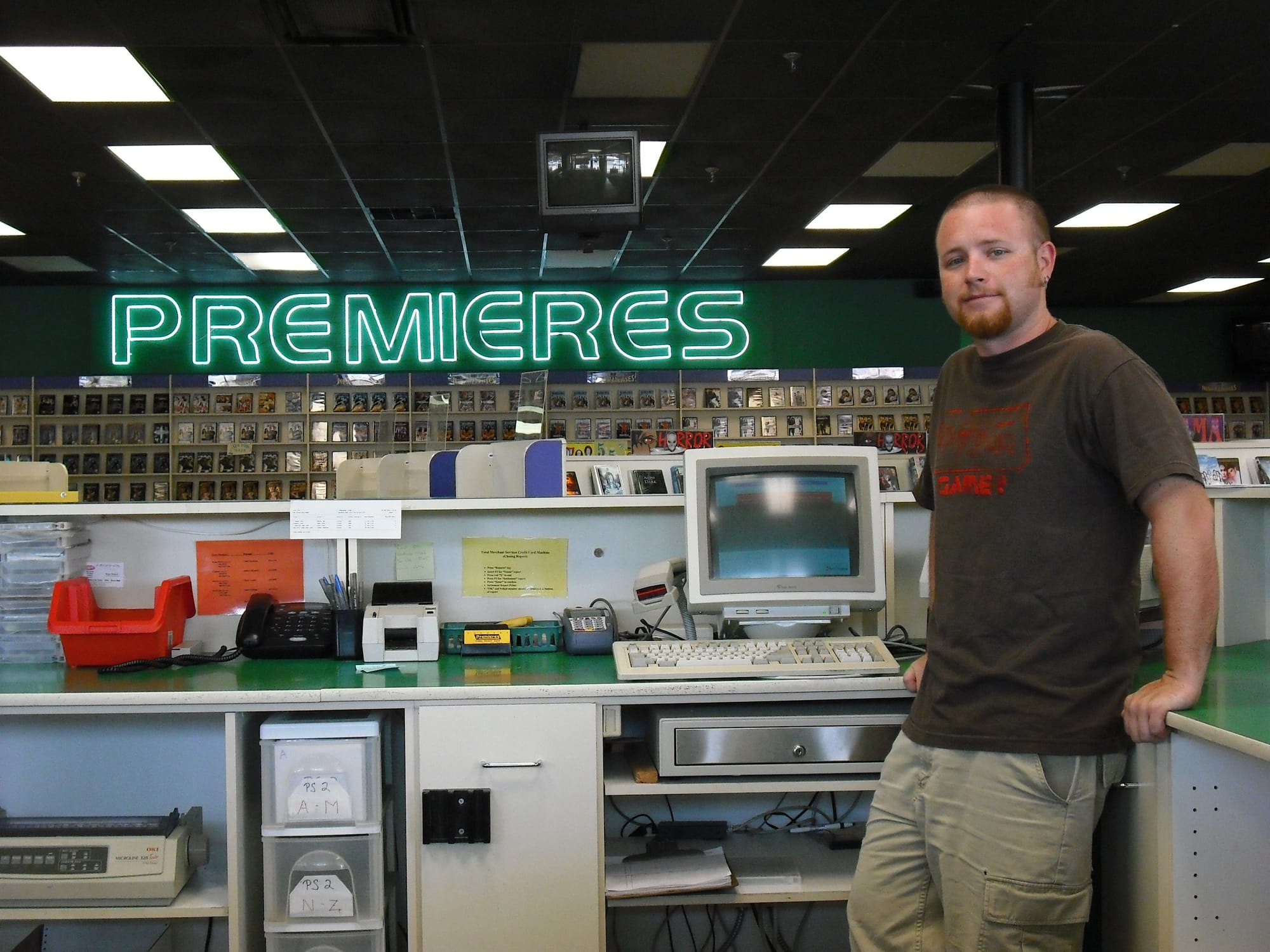 Robert White stands behind the counter at Premieres Video in Clayton, with a large green “PREMIERES” sign, rows of movie rentals, and an old computer system behind him.