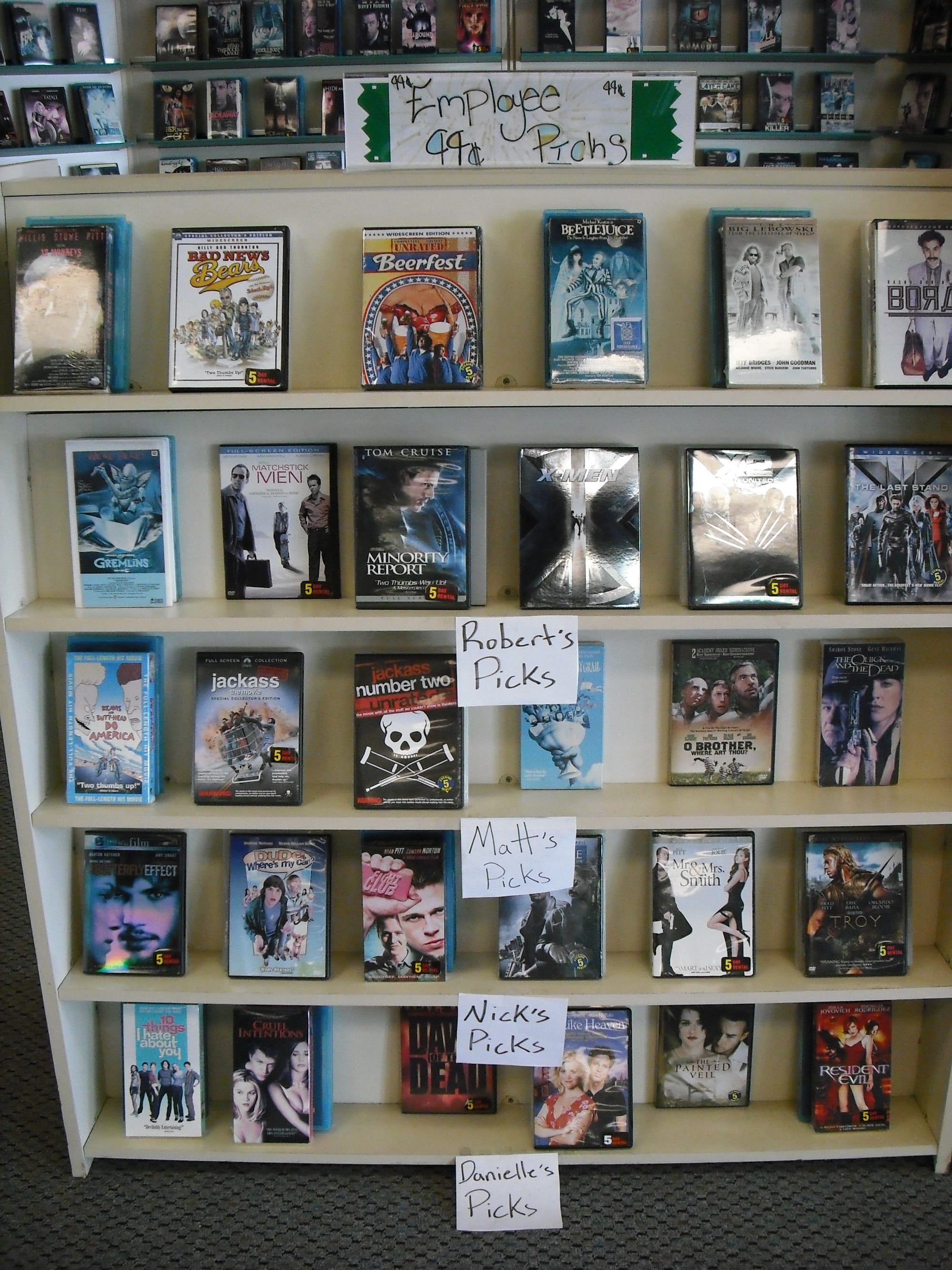 A Premieres Video shelf displays employee 99-cent movie picks, with handwritten signs for Robert’s Picks, Matt’s Picks, Nick’s Picks, and Danielle’s Picks among rows of DVD cases.