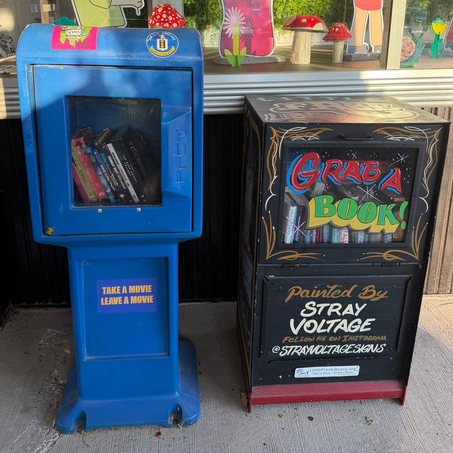 A blue Free Blockbuster box filled with DVDs stands next to a hand-painted black Little Free Library filled with books outside Odd One Out in Downtown Clayton.