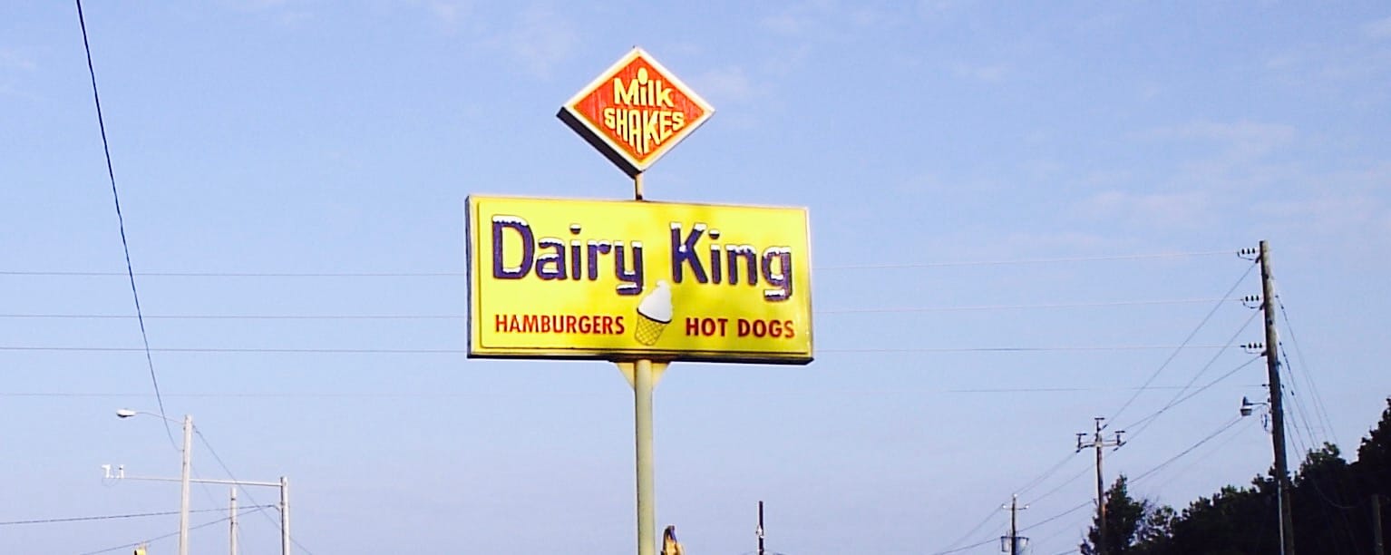 A vintage yellow Dairy King roadside sign advertising milkshakes, hamburgers, and hot dogs against a blue sky, with power lines in the background.