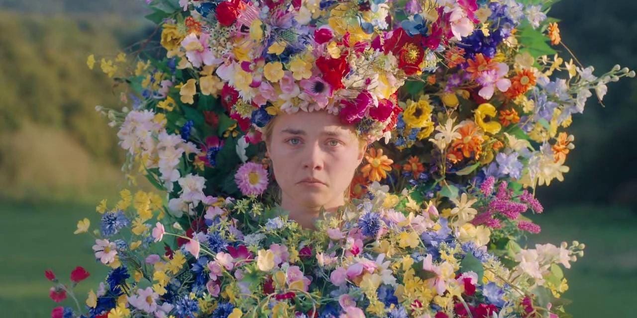 Promotional still from Midsommar showing a woman facing forward while surrounded by an enormous arrangement of brightly colored wildflowers and wearing a large floral crown.