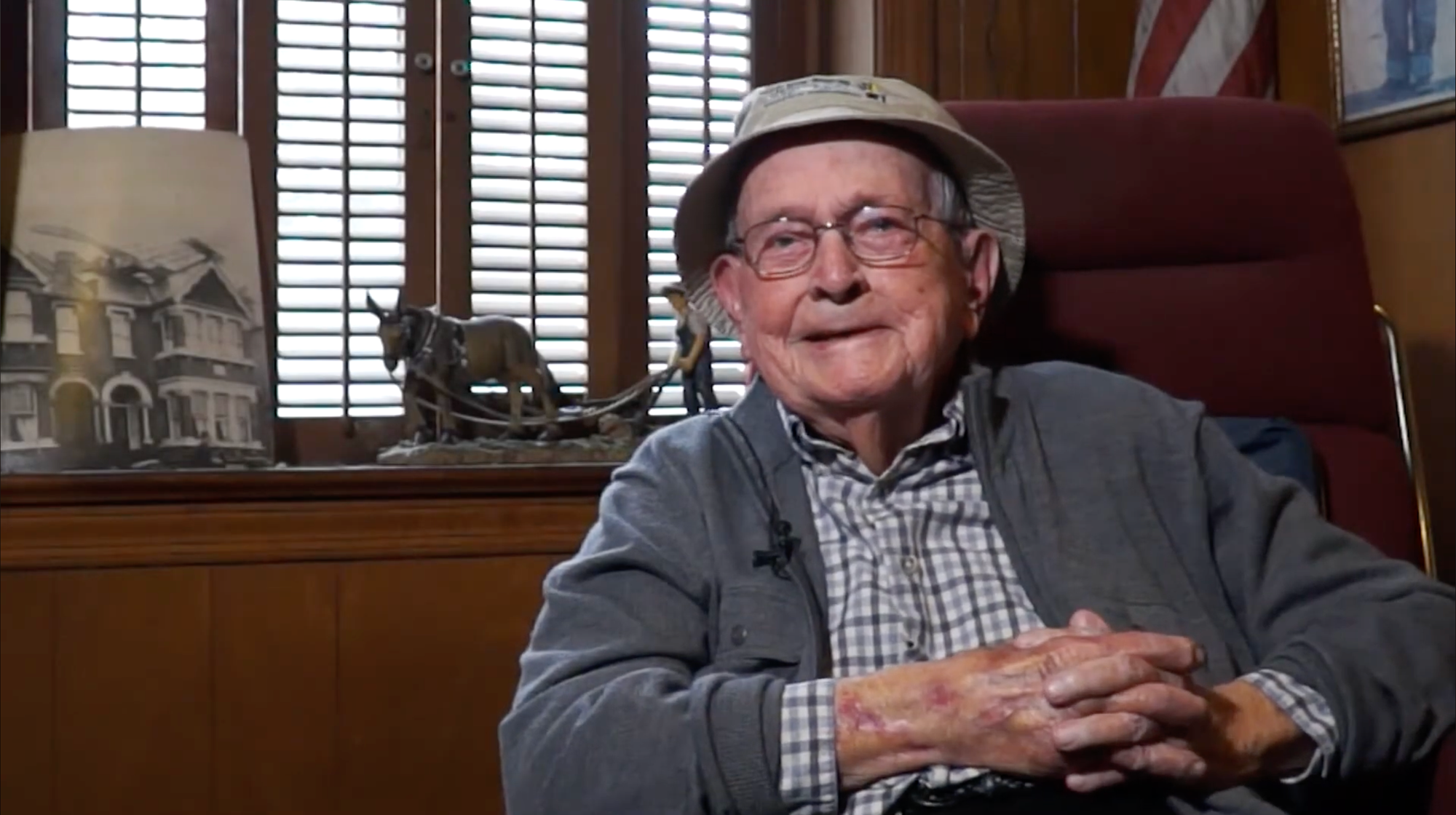Sam Robertson sits in a chair wearing glasses and a light-colored hat, with a model mule team and historic photograph displayed behind him.