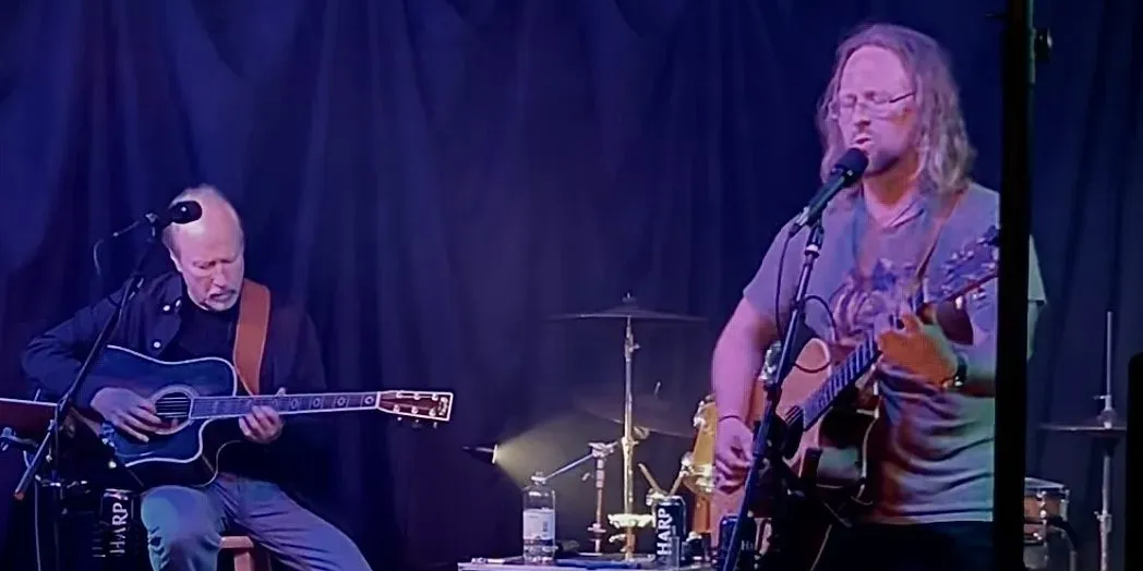 Two musicians perform onstage with acoustic guitars, one seated and one standing at a microphone, with drums and stage equipment in the background.
