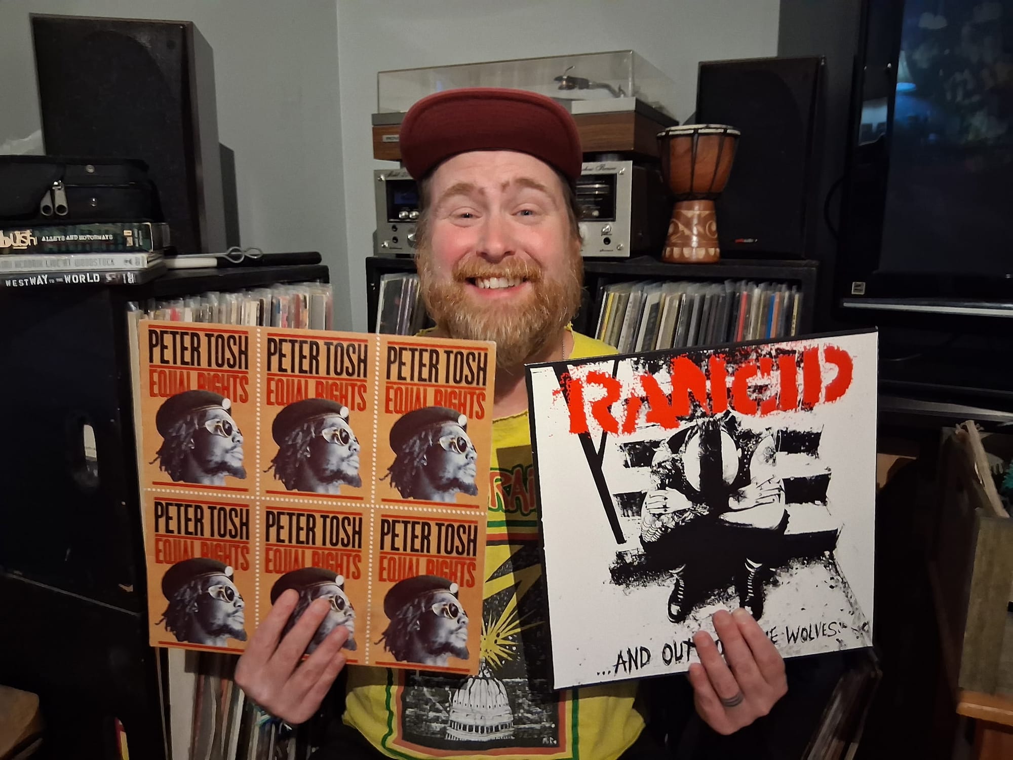 Joel Fitzgerald smiles while holding vinyl copies of Peter Tosh’s Equal Rights and Rancid’s ...And Out Come the Wolves in front of shelves filled with records and stereo equipment.