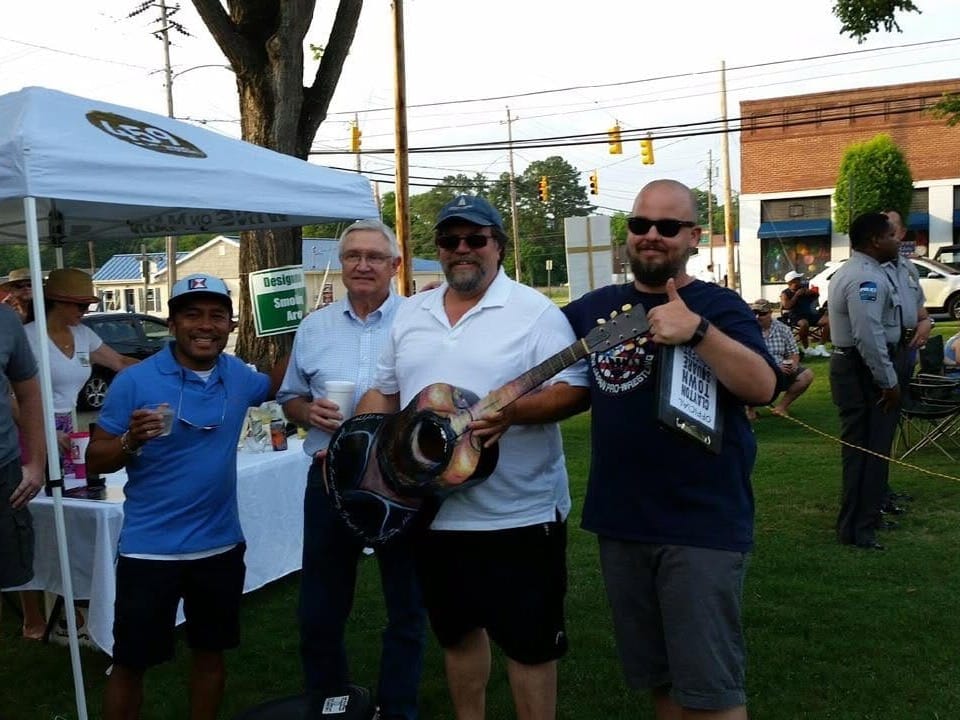 Clark Hipolito, Michael Grannis, Bruce Naegelen and Dan Barbour stand together at an outdoor Downtown Clayton concert as Naegelen holds a hand-painted guitar made by Hipolito.